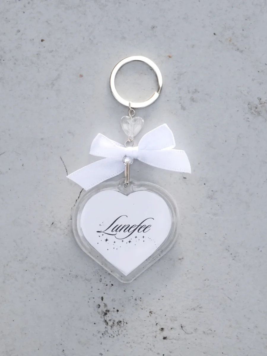 A clear heart keychain with a white ribbon and the Lunefee logo, on grey concrete